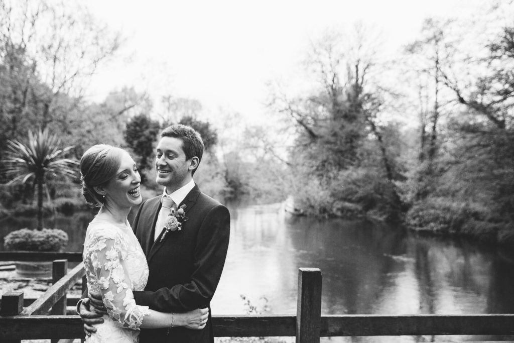 Balck and white image of the bride and groom laughing, standing by the river at their wedding