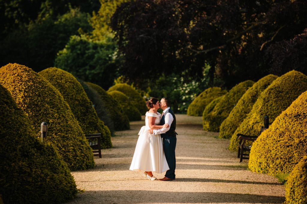 Bride and groom standing by yew trees, manor by the lake