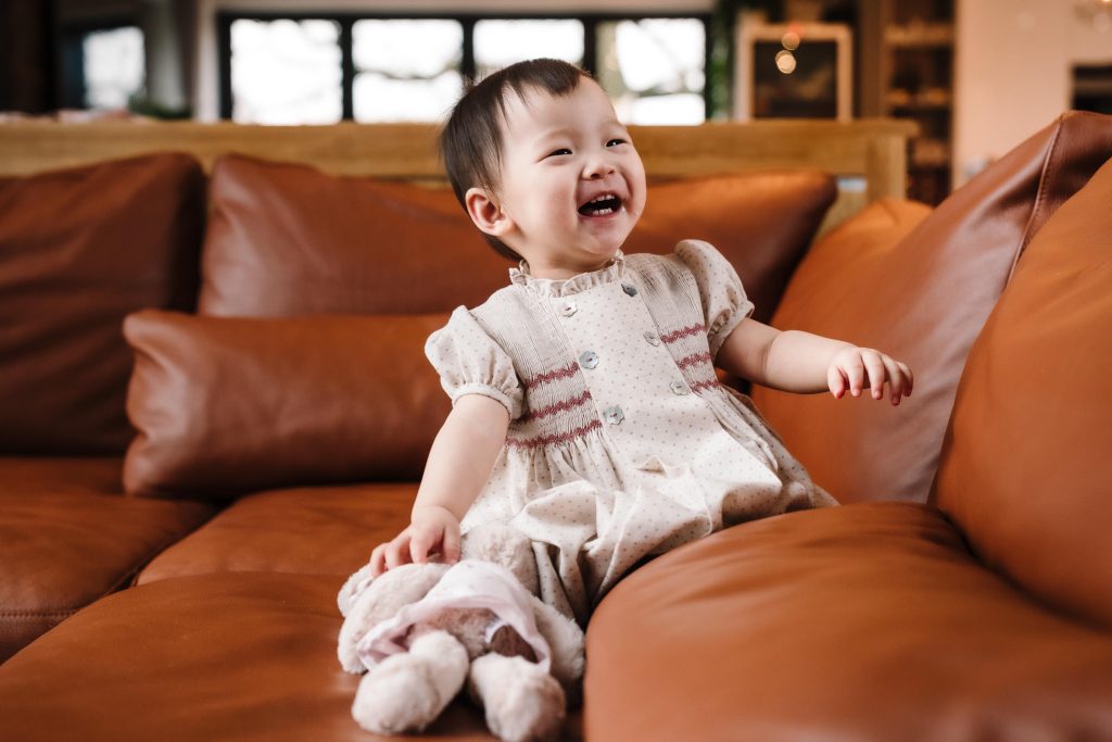 one year old girl laughing on sofa, holding a teddy.  
