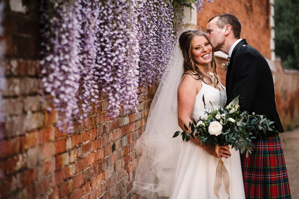 Groom kissing bride against wall with wisteria 
